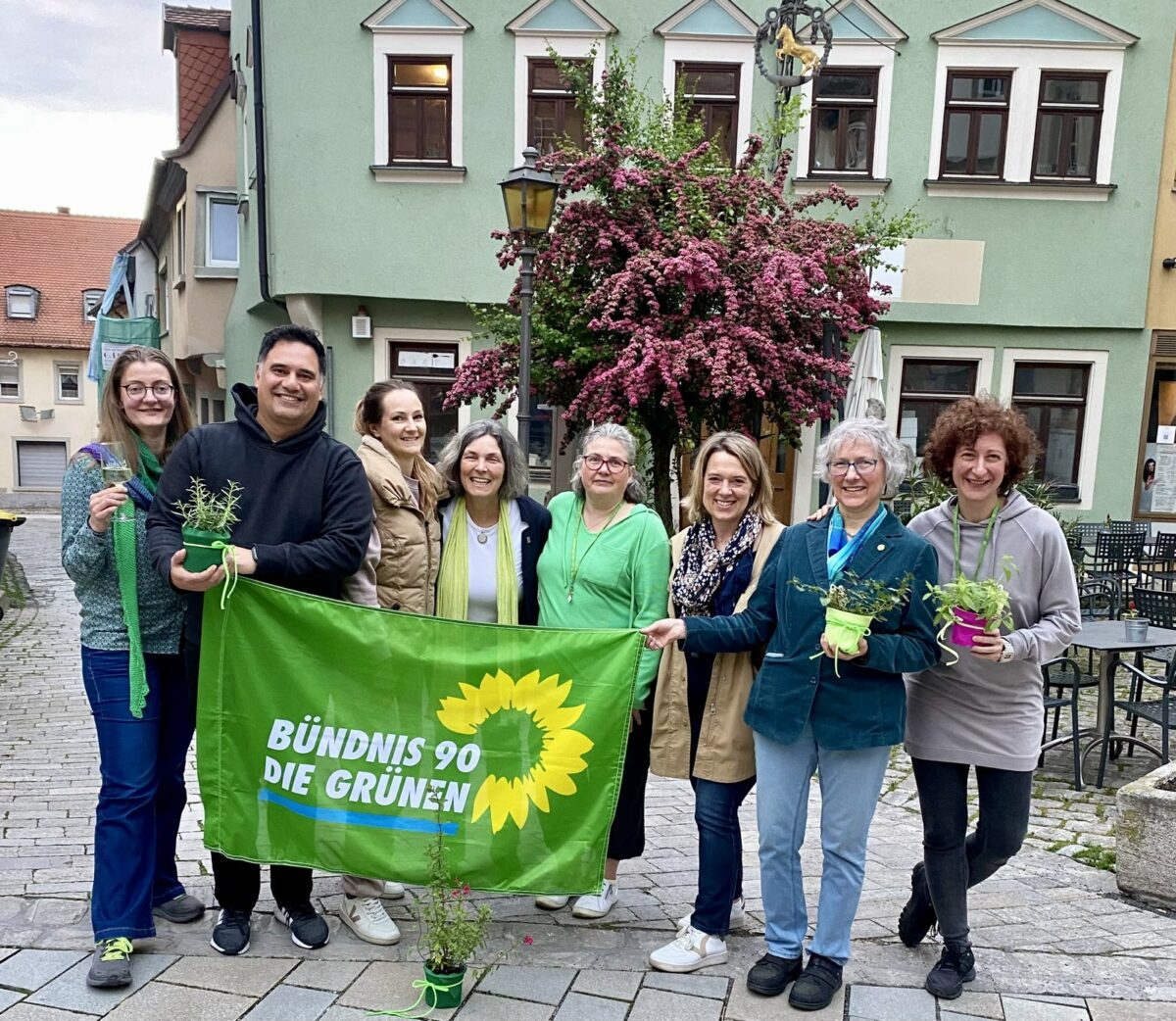 Die unten genannten Personen mit einem grünen "Bündnis 90/Die Grünen" Banner und kleinen Zimmerpflanzen als Geschenk für den Orts-Vorstand in den Händen. Alle lachen in die Kamera. 