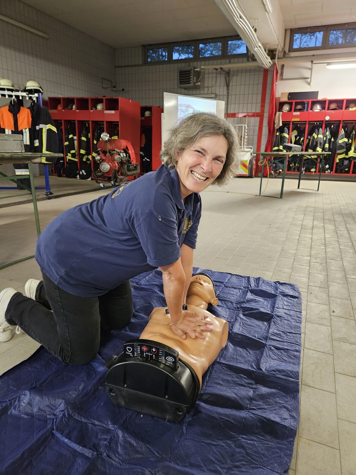 Kerstin Celina sitzt auf dem Boden, vor ihr liegt eine medizinische Trainingspuppe auf der sie simuliert wie erste Hilfe funktioniert. Im Hintergrund zu sehen ist eine Feuerwehrhalle.