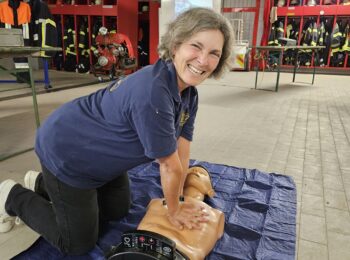 Kerstin Celina sitzt auf dem Boden, vor ihr liegt eine medizinische Trainingspuppe auf der sie simuliert wie erste Hilfe funktioniert. Im Hintergrund zu sehen ist eine Feuerwehrhalle.