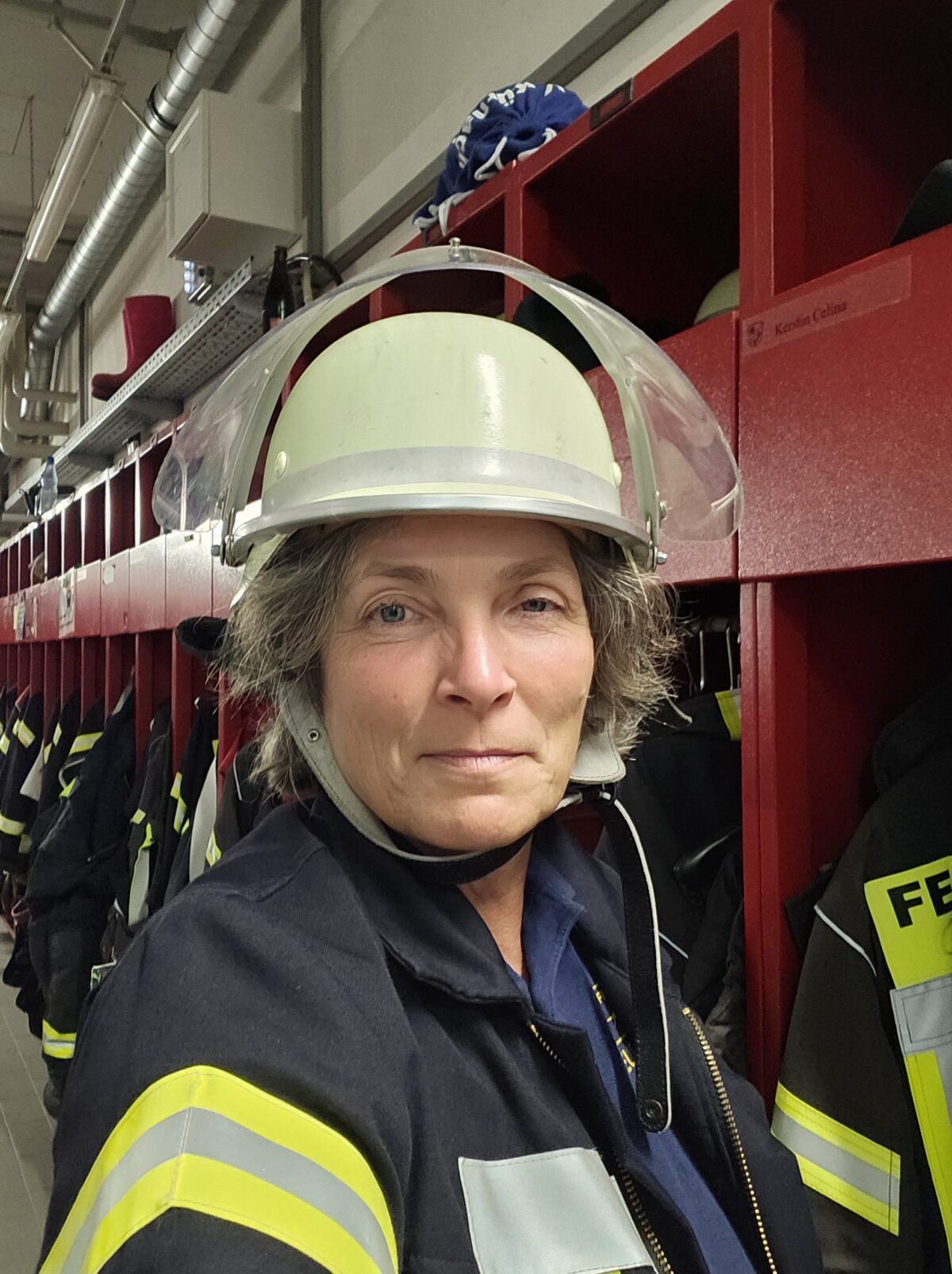 Ein Selfie von Kerstin Celina, die eine Feuerwehrjacke und weißen Helm trägt. Im Hintergrund zu sehen, sind die roten Spinde der Feuerwehr.