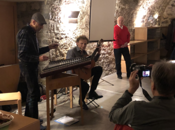 Der Professor und die Musiker stehen vorne vor einer Steinwand. Die Musiker spielen, der Professor schaut zu