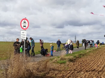 Zu sehen sind eine Reihe von Menschen mit Winddrachen in der Hand und in der Luft. Sie laufen auf einem Weg zwischen Äckern.