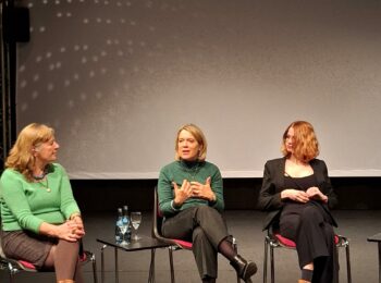 Auf dem Bild sind die Diskussionsteilnehmerinnen Christine Scheel, Jessica Hecht und Lilli Grosch auf einer Bühne zu sehen