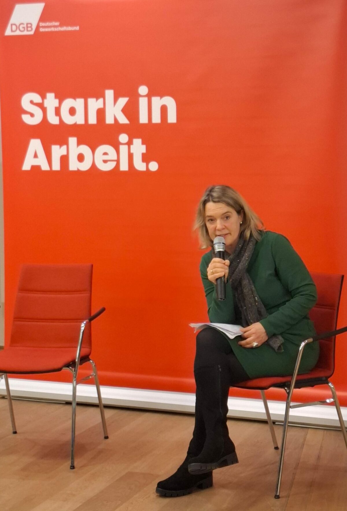 Jessica Hecht beim Heißen Stuhl vom DGB. Sie sitzt vor einer roten Wand auf der "stark in Arbeit" steht. Sie hält ein Mikro in der Hand und spricht gerade