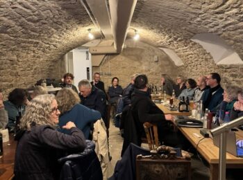 Ein Foto in der Pizzeria. Das Raum ist gewölbt und mit weißen Steinen ausgekleidet. An langen Tischen sitzen Menschen und essen Pizza. Das Bild ist von vorne fotografiert.