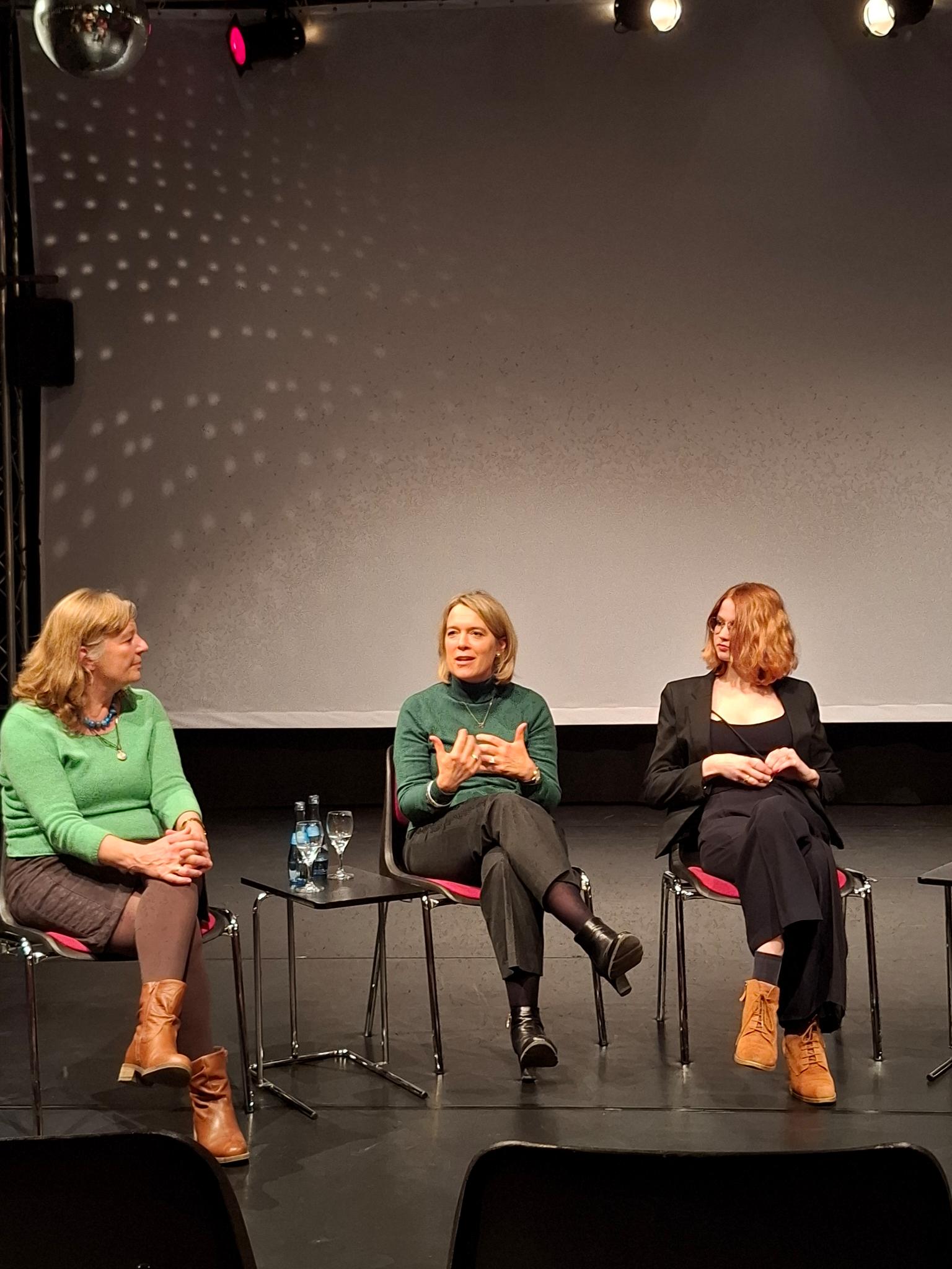 Auf dem Bild sind die Diskussionsteilnehmerinnen Christine Scheel, Jessica Hecht und Lilli Grosch auf einer Bühne zu sehen