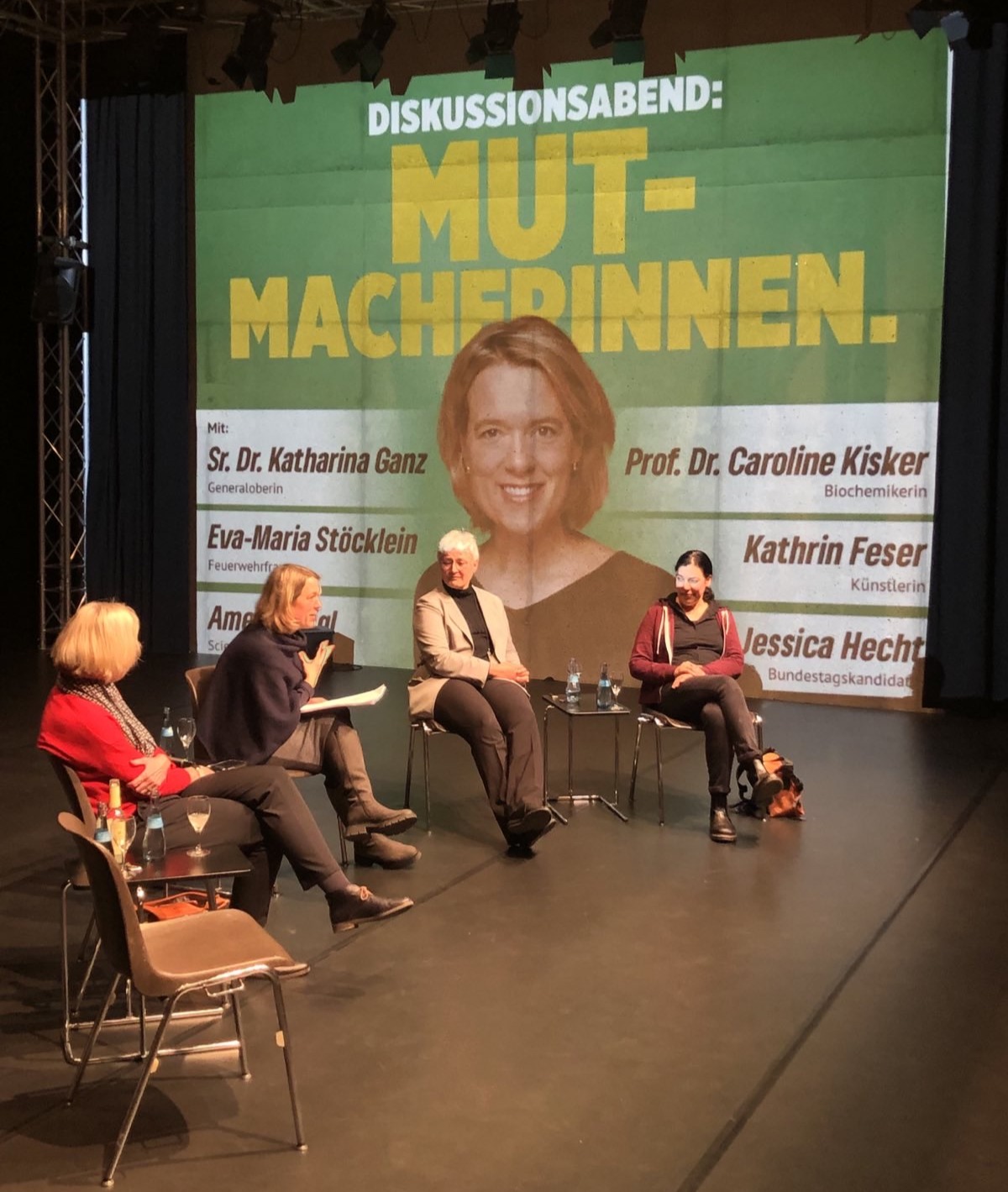 Auf dem Bild ist eine Bühne zu erkennen. Auf dieser Bühne sitzen die Diskussionsteilnehmerinnen. Im Hintergrund ist das Veranstaltungsplakat "Mut-Macherinnen" zu erkennen