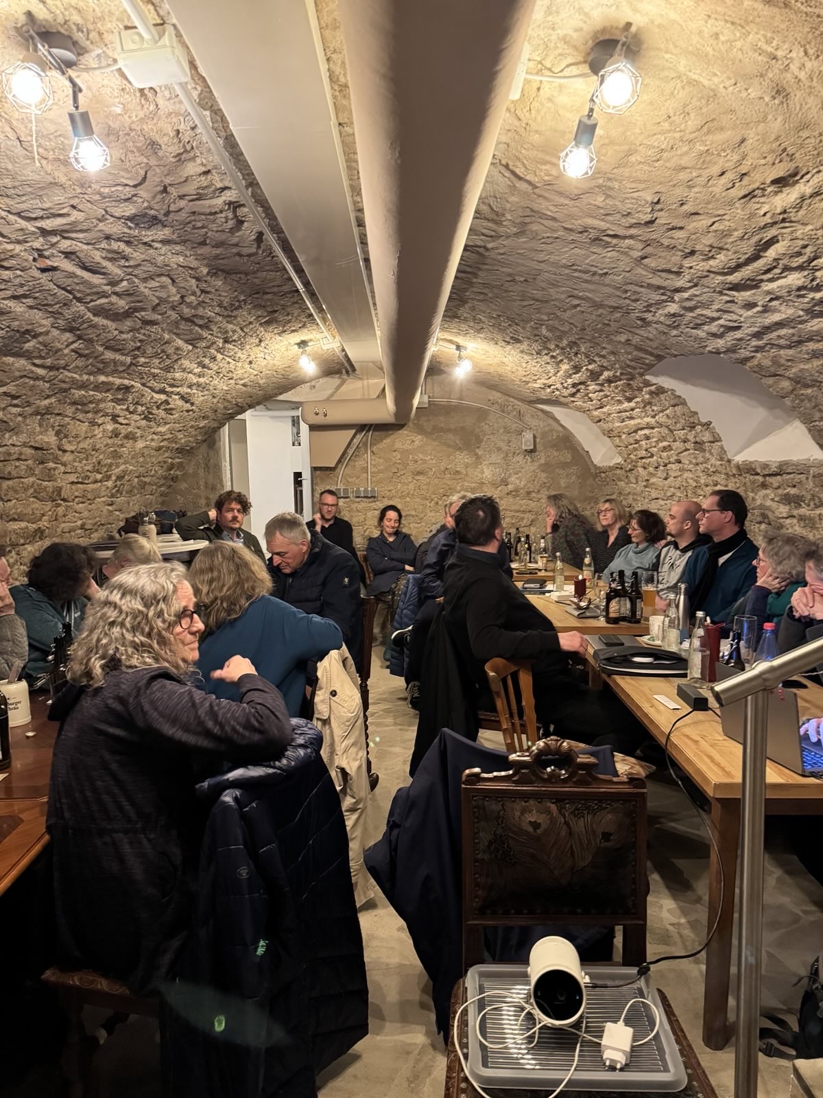 Ein Foto in der Pizzeria. Das Raum ist gewölbt und mit weißen Steinen ausgekleidet. An langen Tischen sitzen Menschen und essen Pizza. Das Bild ist von vorne fotografiert.